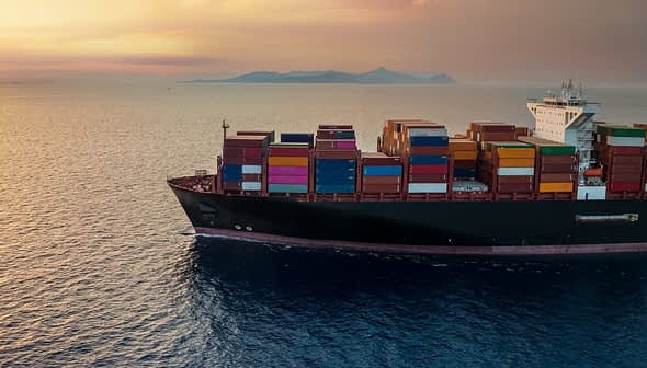 Cargo ship carrying colorful containers sailing on calm waters during sunset. - Olive Oil Times