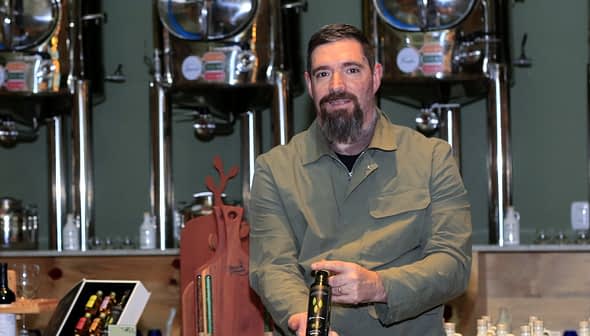 Man with a beard holding a bottle of olive oil in a production facility with metal containers in the background. - Olive Oil Times