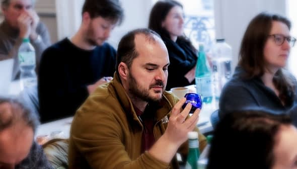 Man examining a blue olive oil bottle while seated in a seminar with other attendees in the background. - Olive Oil Times