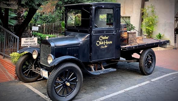 A vintage black pickup truck with wooden barrels in the back, parked on a paved surface. - Olive Oil Times