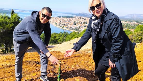 Two individuals bending down to plant a small tree in a rural area with a view of distant hills and water. - Olive Oil Times