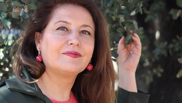 Γυναίκα με μακριά καστανά μαλλιά που φοράει κόκκινα σκουλαρίκια, ποζάρει σε εξωτερικούς χώρους με ένα χέρι σε ένα κλαδί δέντρου. - Olive Oil Times