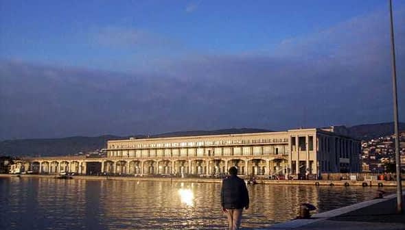Trieste Maritime Station building reflecting on the water during sunset with a person walking nearby. - Olive Oil Times