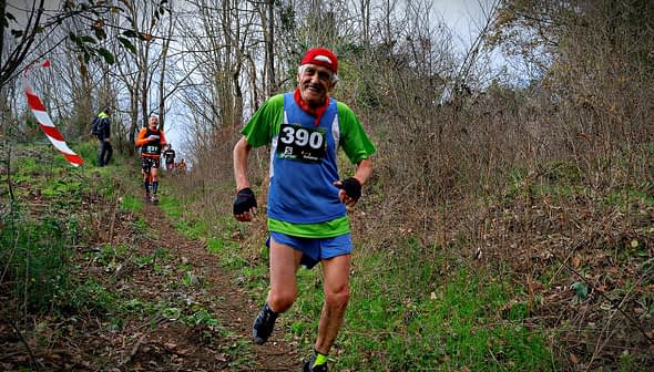 Twee trailrunners doen mee aan een race op een bospad, één draagt ​​een groen-blauwe outfit met een startnummer. Olive Oil Times