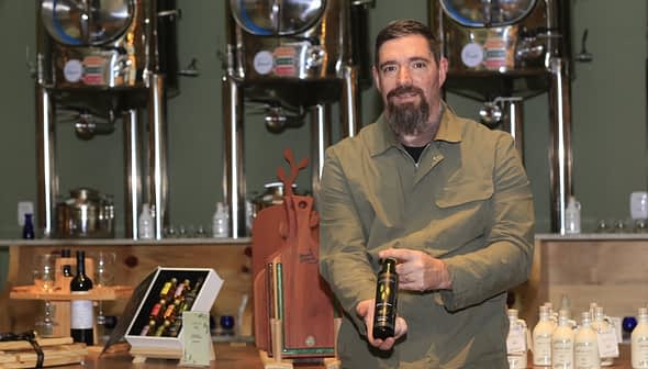 Moški z brado drži steklenico oljčnega olja v proizvodnem okolju z opremo v ozadju. - Olive Oil Times