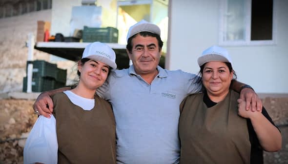 Tri osobe s kapama i radnom odjećom poziraju zajedno na otvorenom. - Olive Oil Times