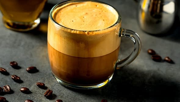 Uma caneca de vidro cheia de café com uma camada distinta de creme no topo, cercada por grãos de café. - Olive Oil Times