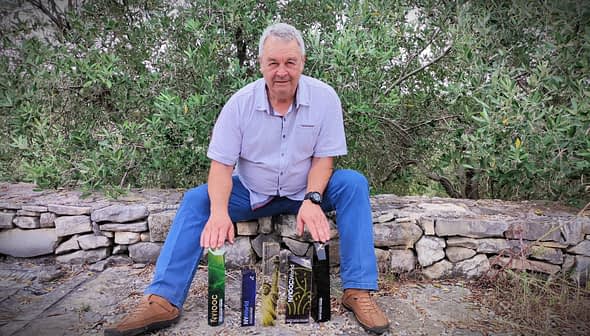 Önünde çeşitli zeytinyağı şişeleri sergilenen taş bir duvarın üzerinde oturan bir adam. Olive Oil Times