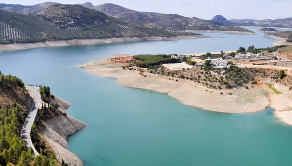 Luftaufnahme eines Stausees mit türkisfarbenem Wasser und Hügeln im Hintergrund. – Olive Oil Times