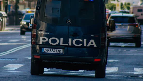 Вид сзади на полицейский фургон с надписью «Policia» на кузове, едущий по городской улице. Olive Oil Times