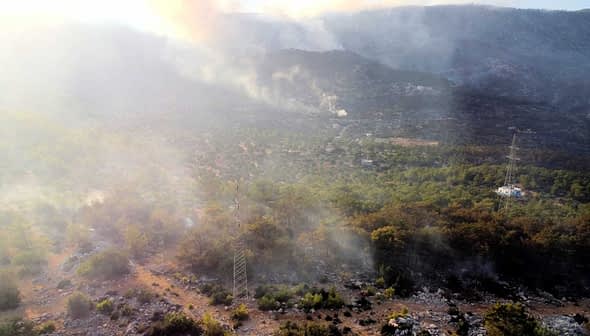 Vista aérea de uma paisagem com fumaça subindo do solo entre árvores e terreno rochoso. - Olive Oil Times