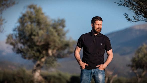 Hombre vestido con un polo negro y pantalones vaqueros de pie al aire libre con olivos al fondo. Olive Oil Times