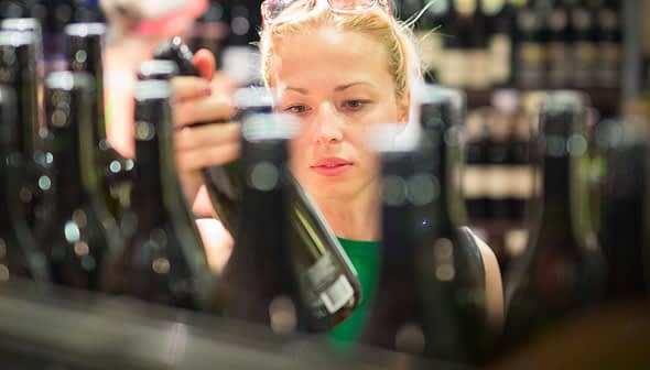 Une femme examine une bouteille de vin lors de ses achats dans un magasin avec plusieurs bouteilles en arrière-plan. - Olive Oil Times