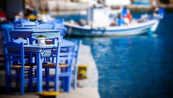Set aus blauen Holzstühlen und einem Tisch mit karierter Tischdecke in der Nähe eines Gewässers. – Olive Oil Times
