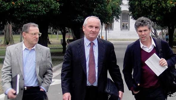 Trois hommes marchant ensemble dans un parc, portant des mallettes et des dossiers. - Olive Oil Times