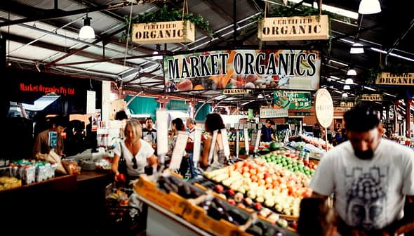Bord met de tekst 'Market Organics' op een drukke boerenmarkt waar verschillende biologische producten worden uitgestald. Olive Oil Times