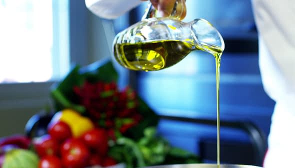 A person pouring olive oil from a glass bottle into a metal bowl in a kitchen setting. - Olive Oil Times