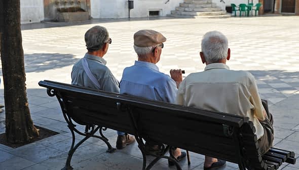 Três homens idosos sentados em um banco, vistos de trás, em uma praça pública. - Olive Oil Times