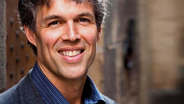 A man with gray hair and a suit smiling at the camera in an outdoor setting. - Olive Oil Times