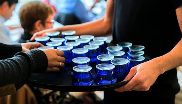 A tray holding multiple blue glasses filled with olive oil being served at an event. - Olive Oil Times