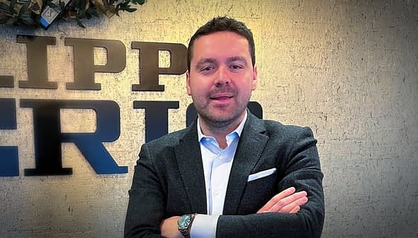 Άνδρας με σκούρο σακάκι και λευκό πουκάμισο που στέκεται με τα χέρια σταυρωμένα μπροστά από έναν τοίχο με υφή. - Olive Oil Times