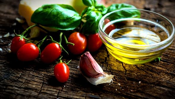 Tomates cereja, alho, manjericão fresco e uma tigela pequena de azeite de oliva em uma superfície de madeira. Olive Oil Times