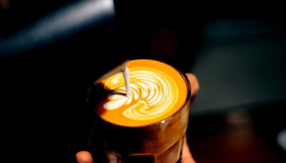 A glass of latte with intricate milk foam art on top, held in a hand against a dark background. - Olive Oil Times