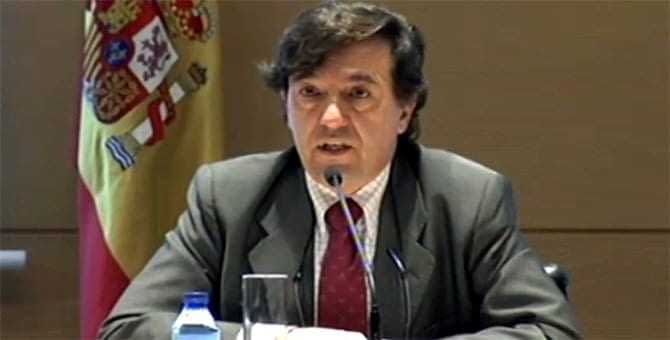 Man in a suit speaking at a conference with a Spanish flag visible in the background. - Olive Oil Times