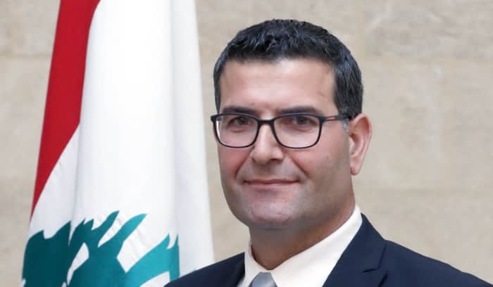 Man wearing glasses and a suit, standing in front of a Lebanese flag. - Olive Oil Times