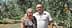 A couple standing together in an olive grove, smiling at the camera. - Olive Oil Times