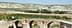 Un pont de pierre à trois arches enjambant une rivière, entouré d'oliviers sur une colline. - Olive Oil Times