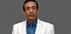 A man wearing a white suit with a blue shirt and tie, standing against a gray background. - Olive Oil Times