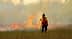 Un pompier en tenue de protection observe un feu de forêt avec des flammes et de la fumée en arrière-plan. - Olive Oil Times