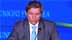 A man in a suit speaking at a podium during a conference with a blue backdrop. - Olive Oil Times