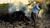 Individual tending to a burning pile of brush in an outdoor setting with smoke rising. - Olive Oil Times