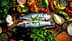 Two whole fish placed on a wooden board surrounded by various vegetables and herbs. - Olive Oil Times