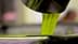 Green olive oil pouring from a metal spout into a container. - Olive Oil Times