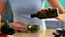 Persona che versa l'olio d'oliva da una bottiglia di vetro scuro su un'insalata fresca in una ciotola trasparente. Olive Oil Times