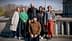 A group of six individuals posing together outdoors near a body of water. - Olive Oil Times