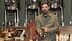 Man with a beard holding a bottle of olive oil in a production setting with equipment in the background. - Olive Oil Times