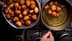 Eine Schüssel gefüllt mit goldbraun frittierten Teigbällchen neben einer Bratpfanne, in der weitere Teigbällchen abgetropft werden. - Olive Oil Times