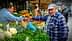 Um homem de boné azul interage com um vendedor em um mercado de vegetais repleto de diversos produtos frescos. Olive Oil Times