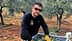 Un homme portant des lunettes de soleil et des gants trie des feuilles d'olivier dans un conteneur dans une oliveraie. - Olive Oil Times