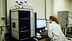 Laboratory technician in a white coat operating analytical equipment and reviewing data on a computer screen. - Olive Oil Times