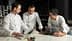 Tres chefs con batas blancas examinan un libro de cocina mientras discuten presentaciones culinarias. Olive Oil Times