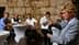 A woman in a gray blazer pouring olive oil while a group of men observes at a tasting event. - Olive Oil Times