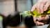 Een persoon schenkt olijfolie uit een glazen fles met een groene dop, waarbij olie uit de tuit druppelt. Olive Oil Times