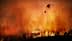 Un hélicoptère largue de l'eau sur un grand incendie de forêt avec des flammes et de la fumée en arrière-plan. - Olive Oil Times