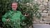 Hombre con una chaqueta verde de pie entre ramas de olivo con aceitunas en la mano. - Olive Oil Times