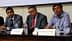 Three men seated at a conference table with water bottles and nameplates in front of them. - Olive Oil Times